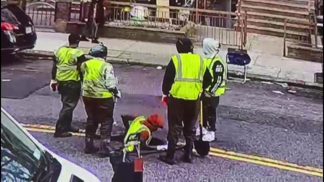 Four men wearing neon yellow vests stand around a manhole in the middle of the street while a fifth man in a yellow vest climbs down into the manhole. 